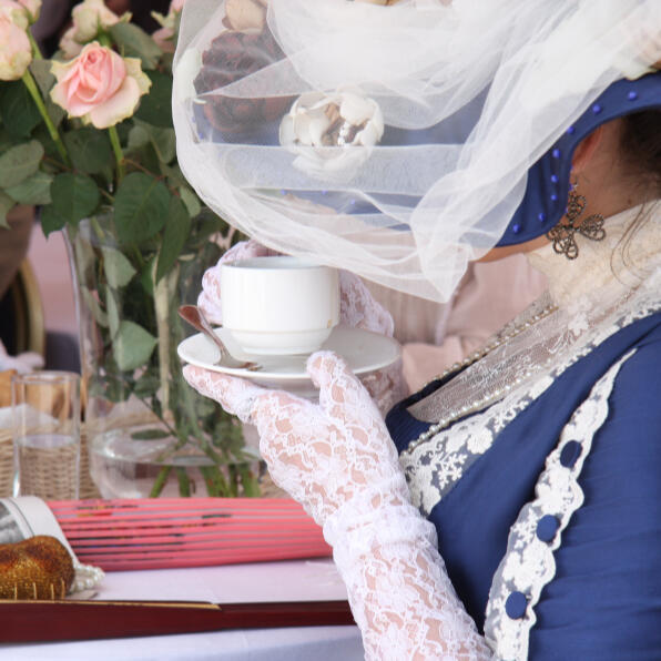 An image of a woman wearing a blue victorian gown with white lace, drinking tea. she is wearing a matching hat with netting that obscures her face.