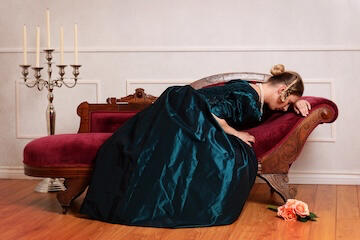 An upset Victorian woman in a green gown resting her head against a burgundy sofa.